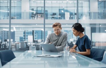 An image for a blog on "is now a good time to invest" image is cgi of two people sat in a conference room , male on left , female on right both looking at a pc with some documents and two glasses of water on the table